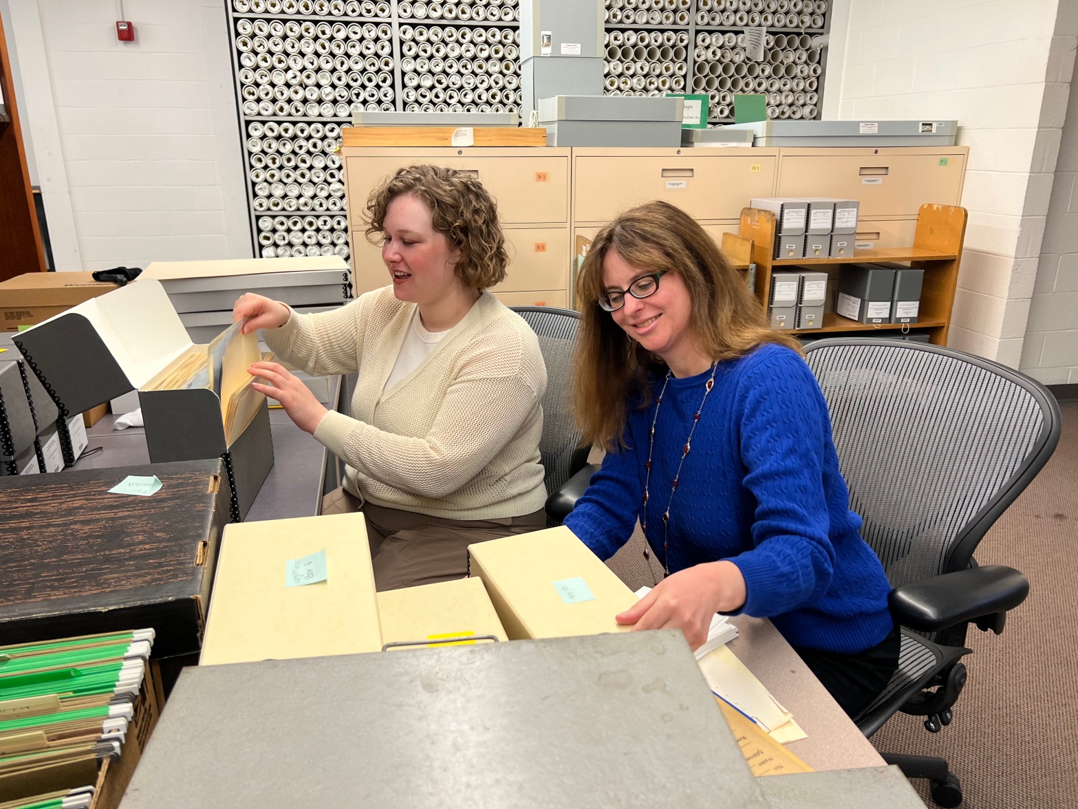 Two women sit at a desk. One is pulling a folder from a document box. The other is looking down at materials on the desk.